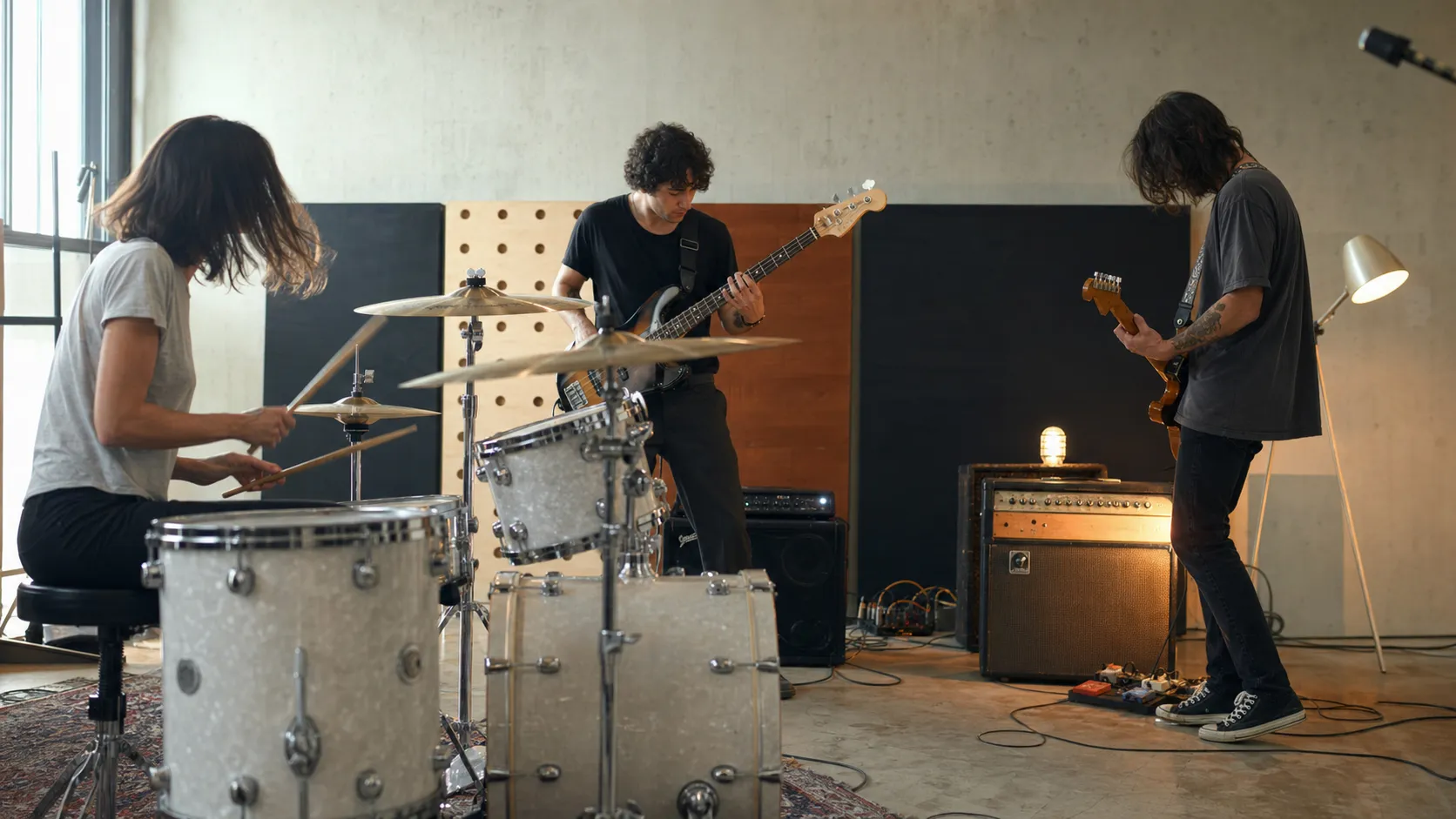 An anonymous drummer, bassist, and guitarist rehearse together in a bright contemporary live room.